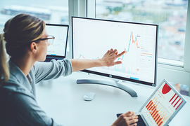 A person pointing at a rising graph on a computer screen, with additional charts displayed on a tablet and another monitor.