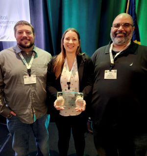 A group photo of three people from Team A in Kentucky, standing together at an event. The person in the center is holding an award and all are wearing event badges.