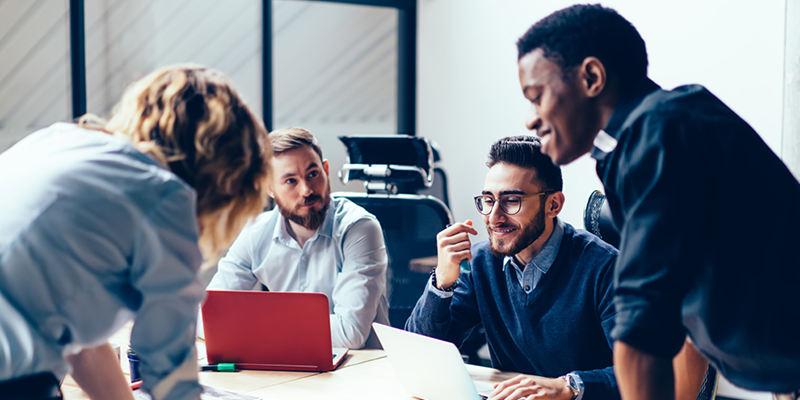 A group of diverse professionals collaborating around a table in a modern office setting, using laptops and discussing ideas.
