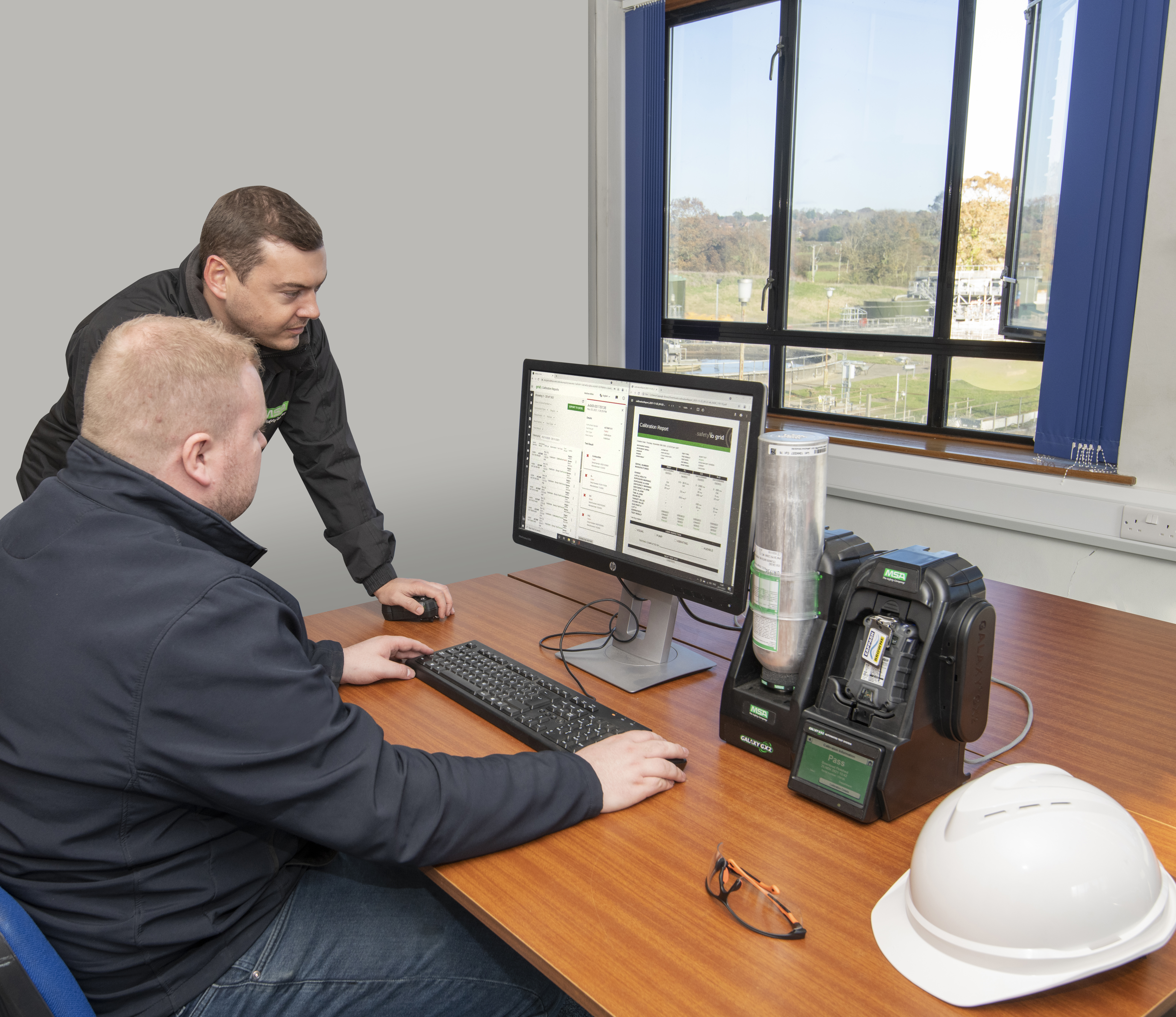 Two technicians are working at a desk in an office, monitoring data on a computer screen. There is industrial equipment on the desk, and a white safety helmet and glasses are also present. A window shows an outdoor industrial facility in the background.