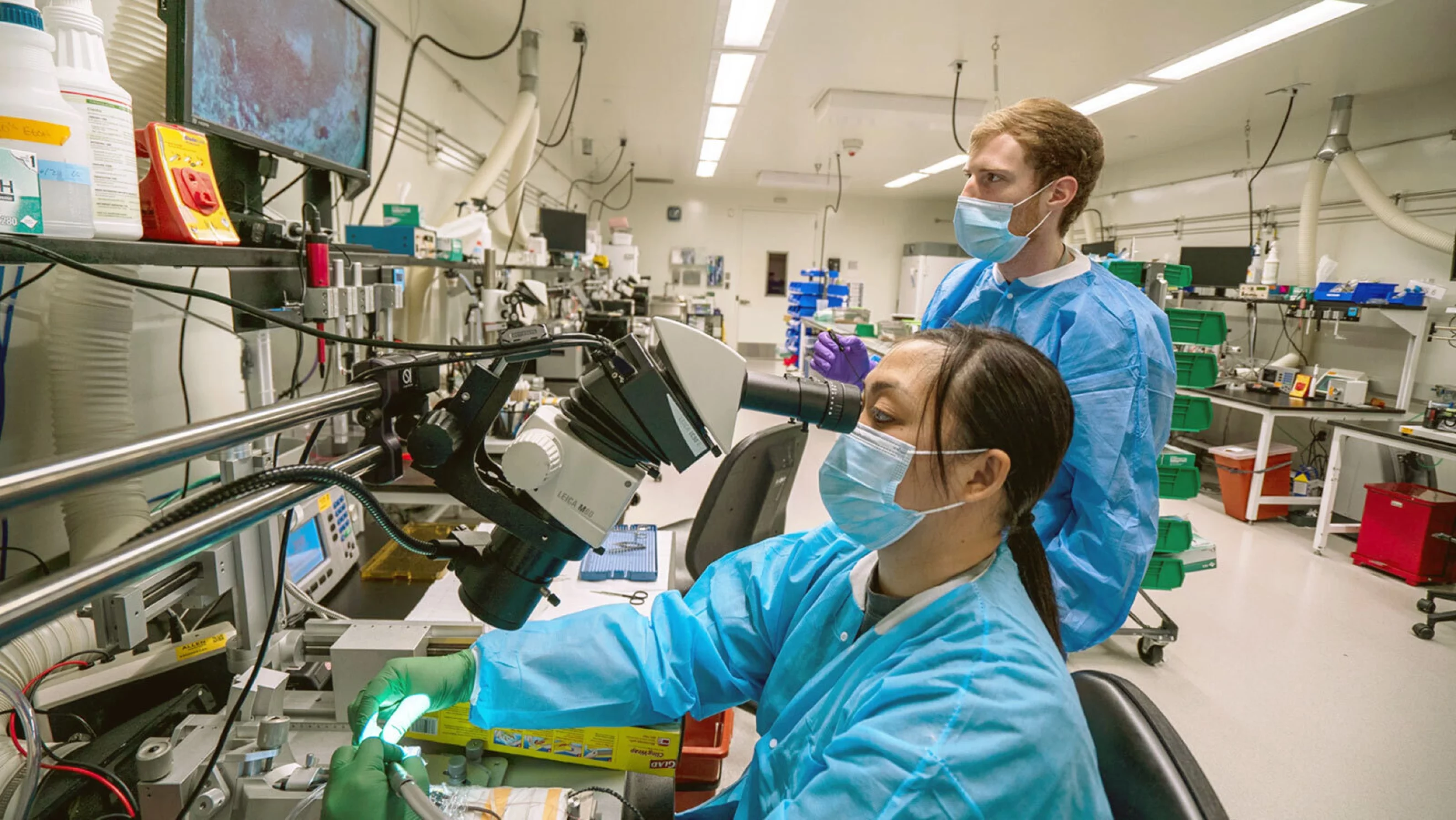Two scientists at the Allen Institute working in a laboratory, wearing masks and blue lab coats. One scientist is using a microscope while the other observes, surrounded by laboratory equipment and monitors.