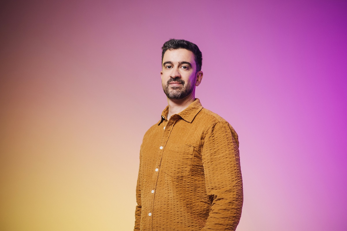 A man wearing a brown textured shirt poses against a soft yellow-to-purple gradient background in this studio portrait.