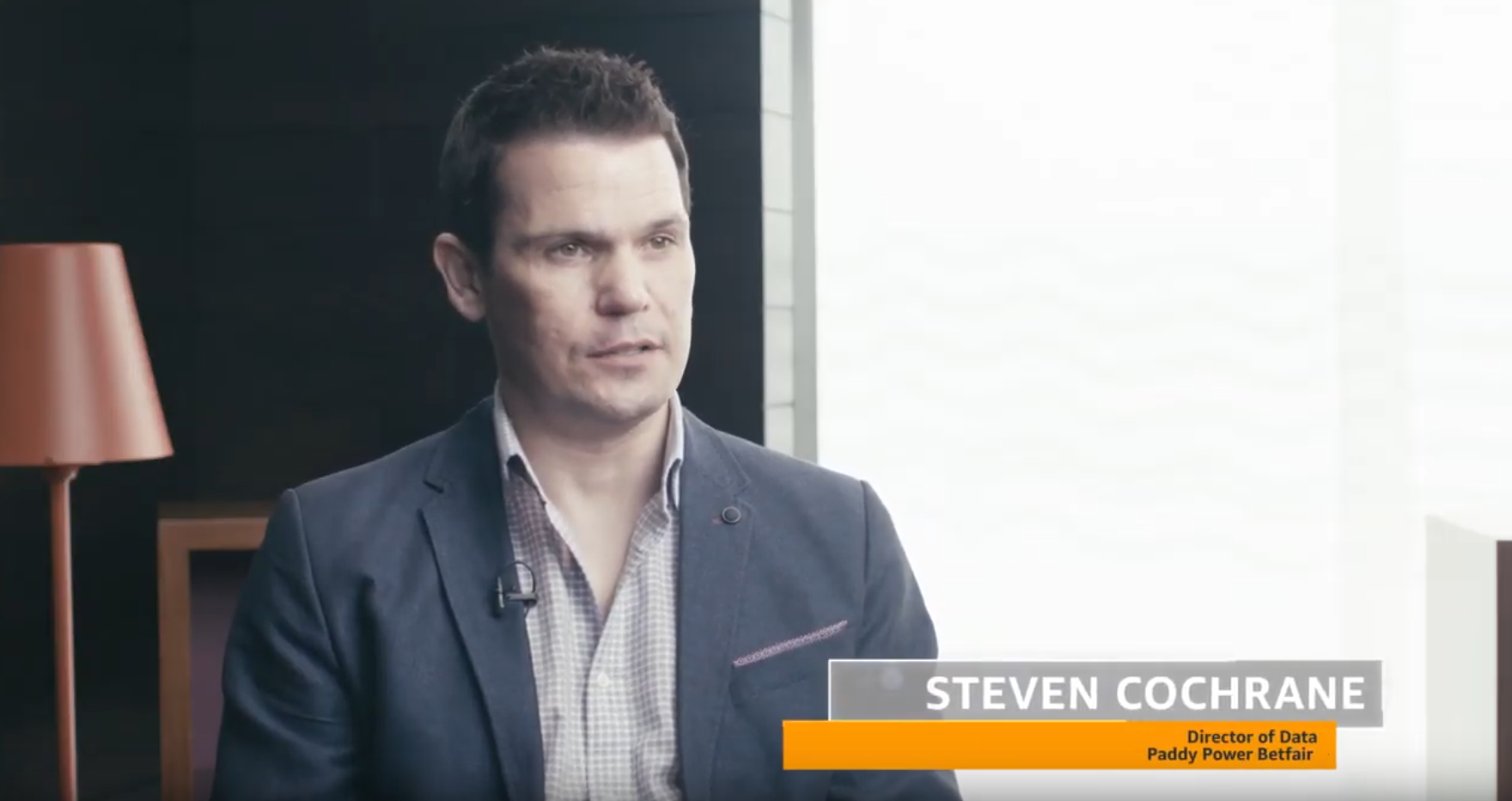 A man in a suit is seated indoors, speaking in an interview setting. Text on the image identifies him as Steven Cochrane, Director of Data at Paddy Power Betfair. There is a lamp and modern office decor in the background.