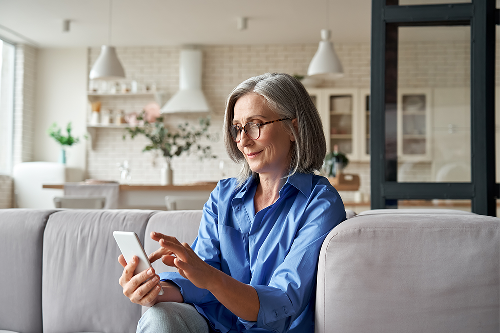 A smiling senior woman with gray hair, wearing glasses and a blue shirt, is sitting on a couch at home and using a smartphone. The background features a modern kitchen and living space with natural light.
