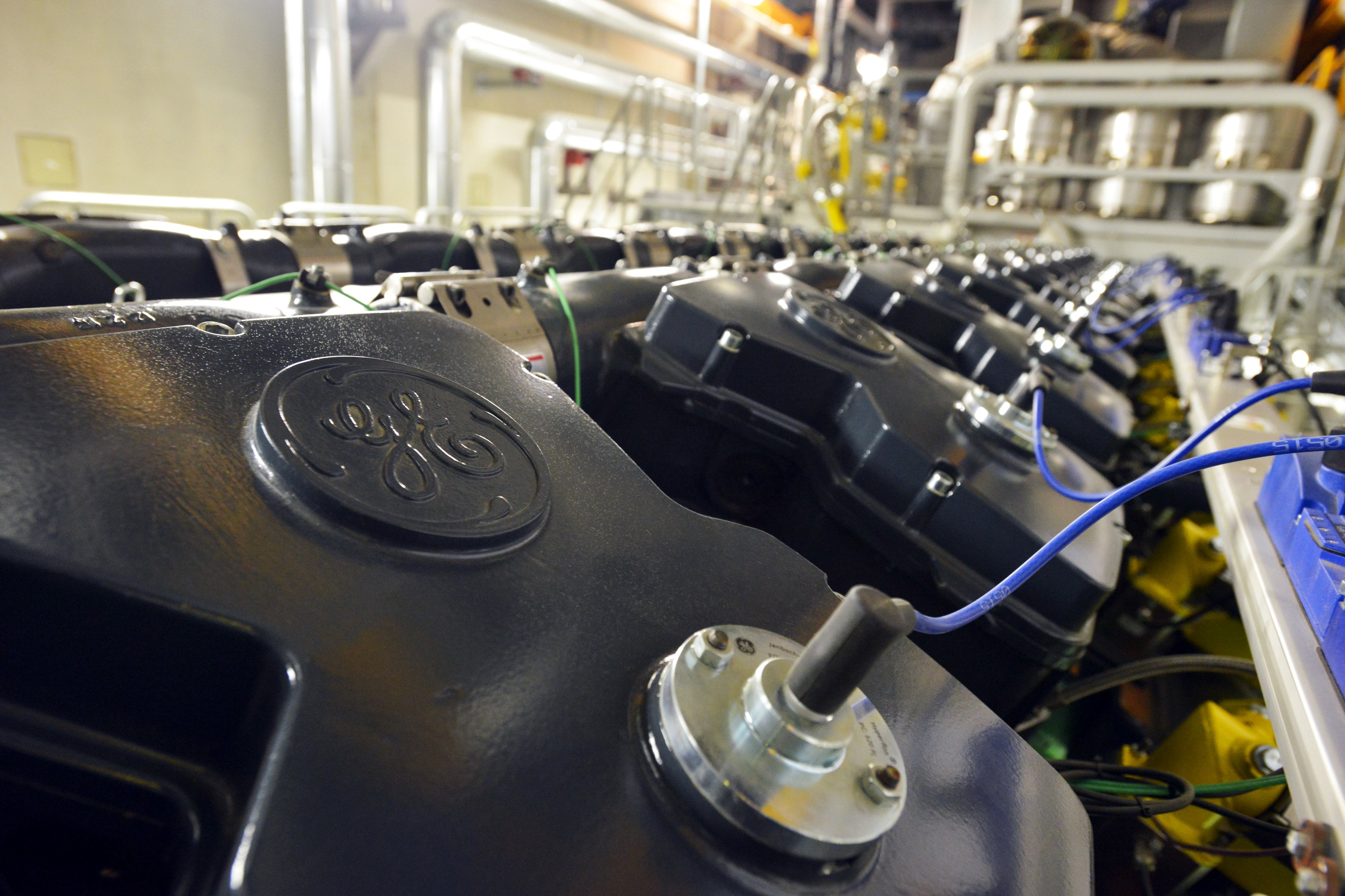 A detailed close-up image of industrial machinery showing a row of black engine components with the GE (General Electric) logo, blue wiring, and stainless steel piping in an industrial facility.