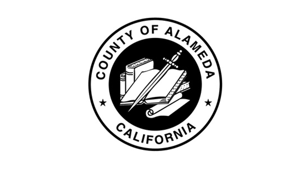 The official seal of Alameda County, California, featuring a black and white circular emblem with books, a sword, and a scroll in the center, surrounded by the words 'County of Alameda California.'