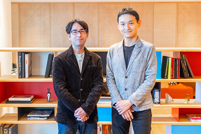 Two individuals standing in front of a modern bookshelf filled with books and decorative items.
