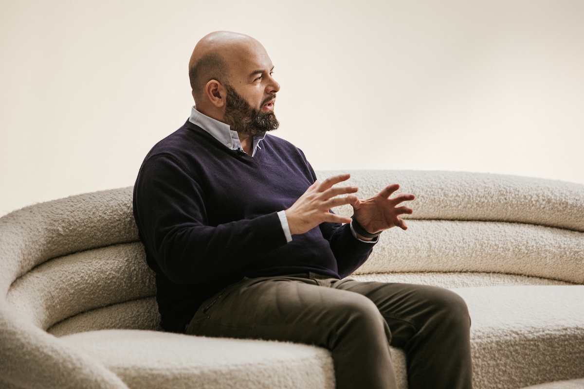 A person seated on a textured curved sofa gestures with their hands while engaged in conversation, set against a plain light background.