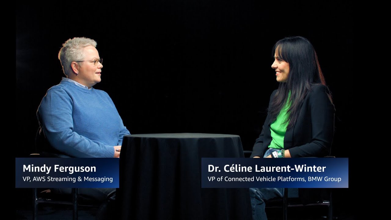 Two individuals seated at a round table in a dark setting, with nameplates identifying them as Mindy Ferguson, VP of AWS Streaming & Messaging, and Dr. Céline Laurent-Winter, VP of Connected Vehicle Platforms, BMW Group.