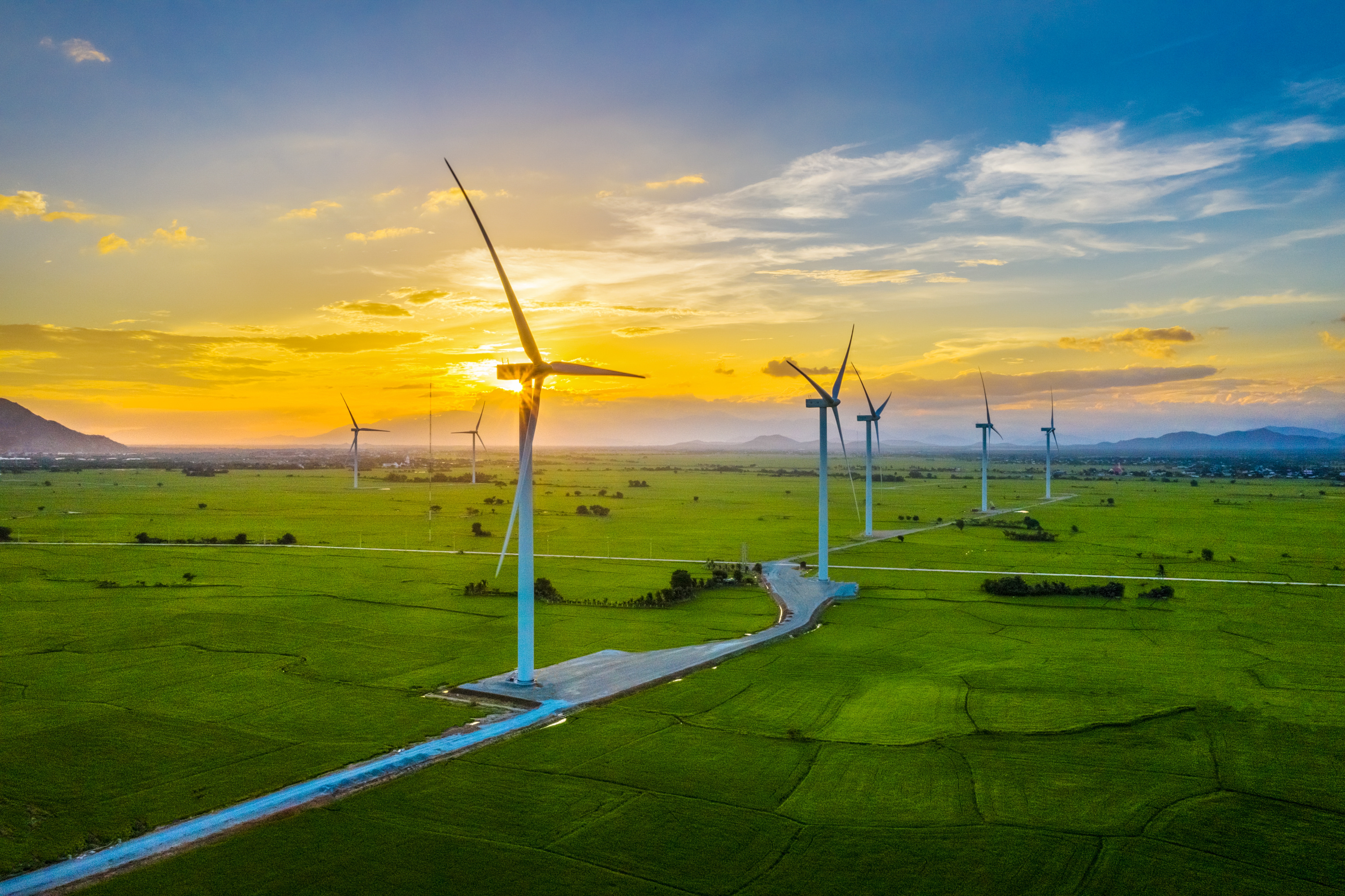 A scenic view of wind turbines on a green field during sunset, generating renewable energy. The sky is bright with orange and blue hues, and mountains appear in the background.