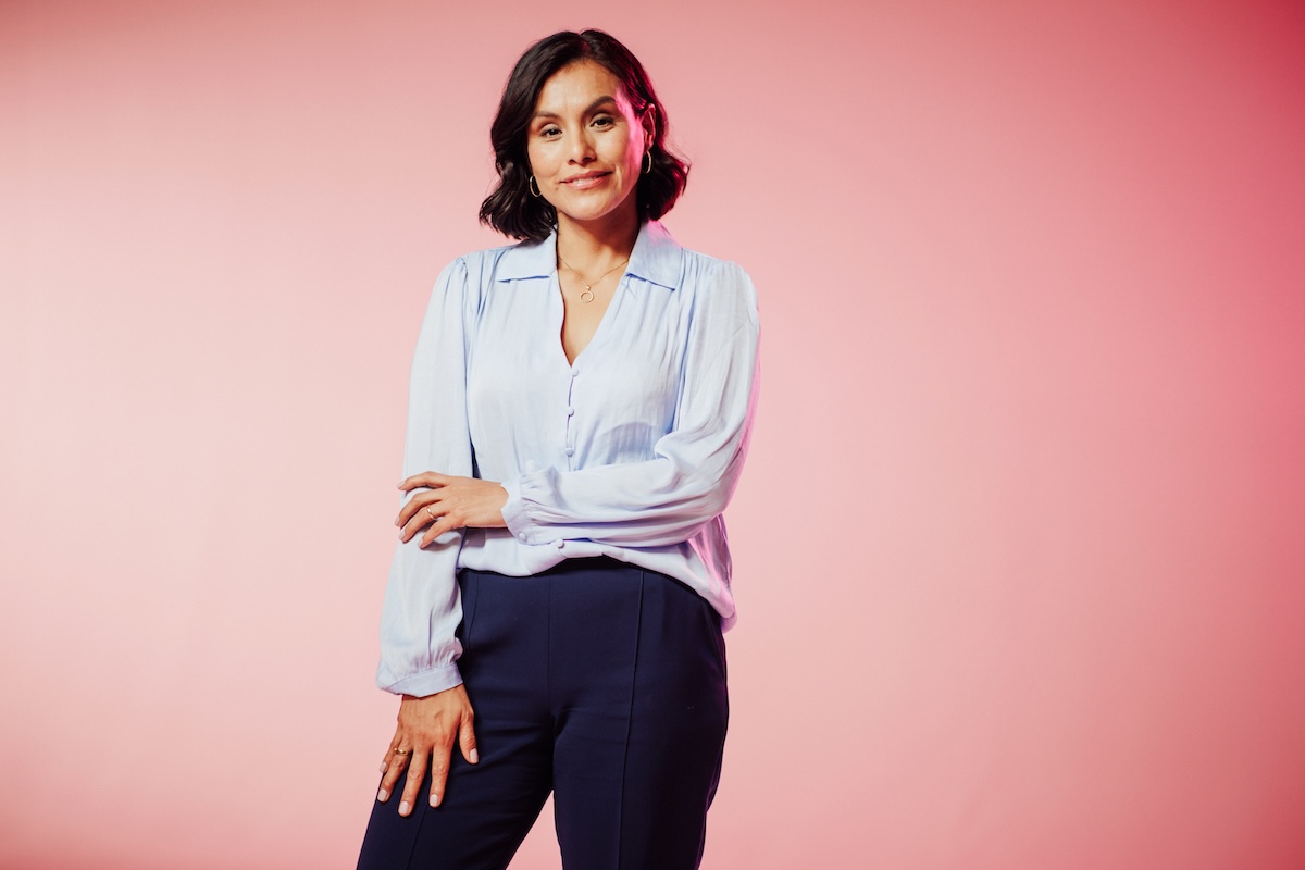 Portrait of a woman wearing a light blue blouse and dark pants standing against a pink background.