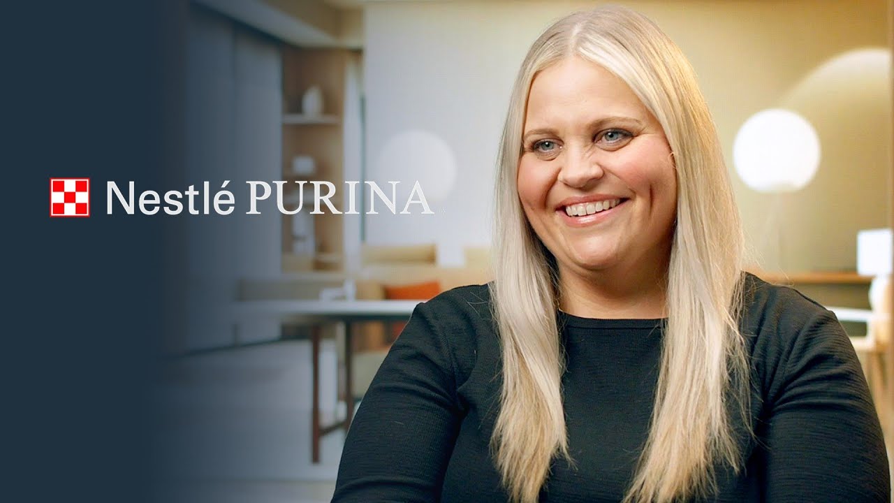 A smiling woman with long blonde hair in a black top is seated in a well-lit office environment. The Nestlé Purina logo is displayed on the left side of the image.