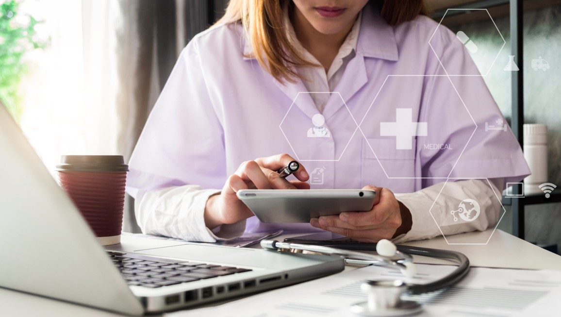 A healthcare professional uses a tablet and laptop at a desk, with medical icons and a stethoscope visible, representing digital health and medical technology.
