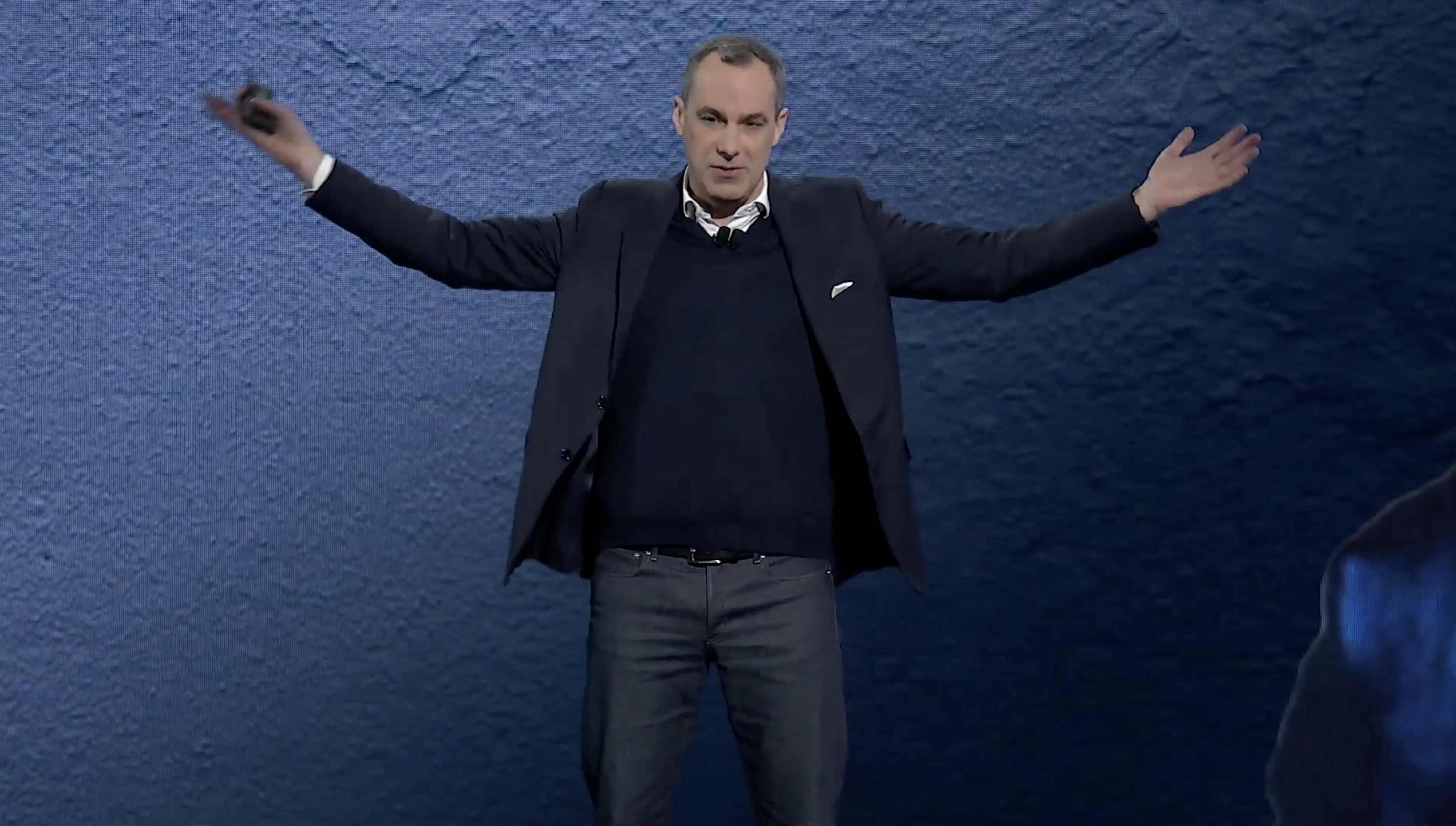 A person in a suit and sweater stands with arms outstretched against a textured blue background.