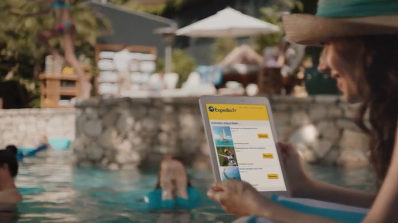 A person relaxing by a pool using a tablet to browse the Expedia website, demonstrating leisure travel and technology usage. The image highlights AWS agility and resiliency in a hospitality setting.