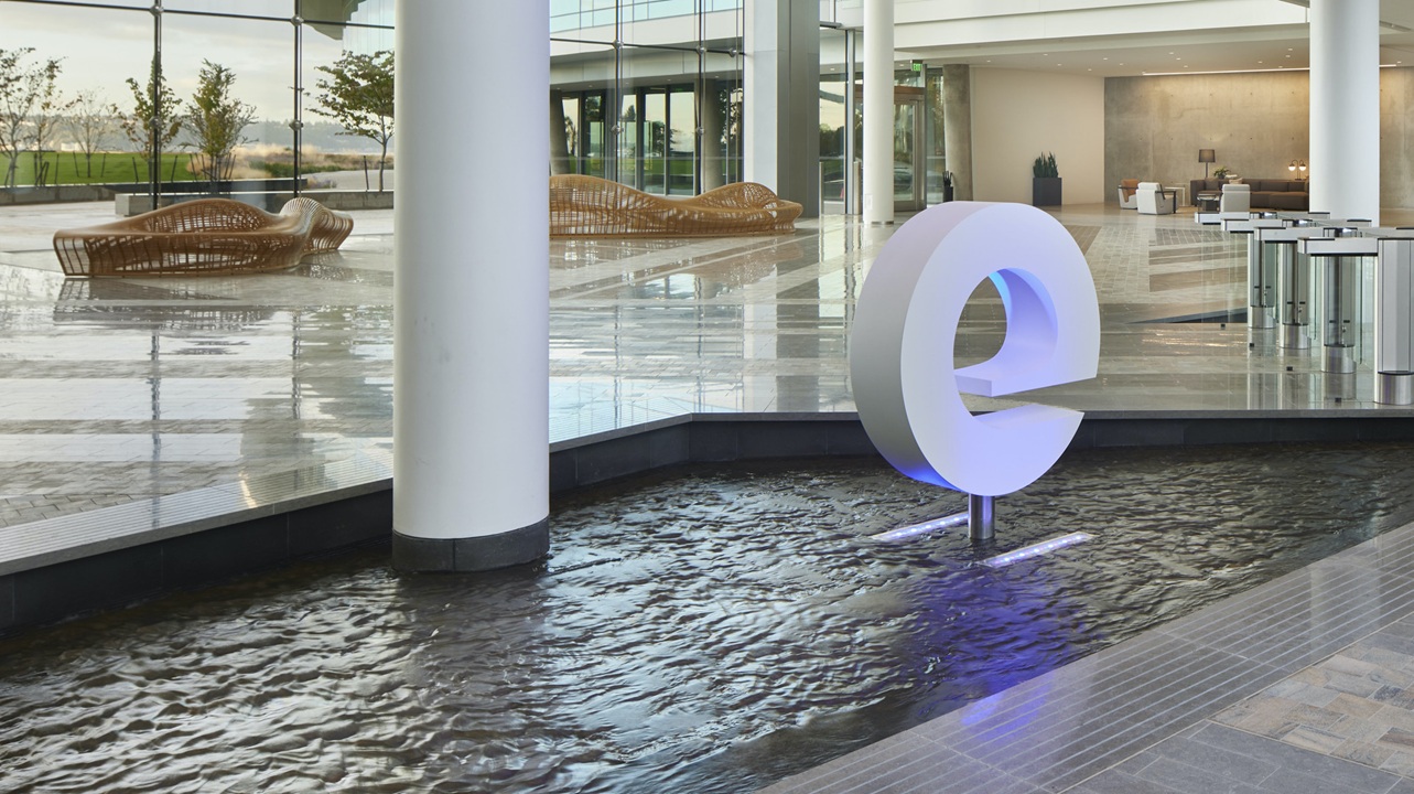 The lobby of Expedia's headquarters featuring a modern sculpture of the letter 'e' illuminated above a water feature, with contemporary seating and open architecture in the background.