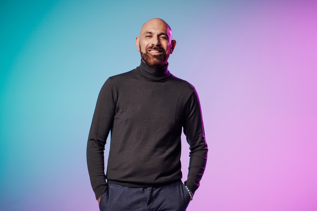 A person wearing a black turtleneck and navy pants stands against a gradient background of blue and pink.