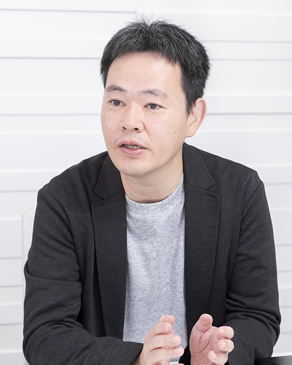 A person wearing a gray shirt and black blazer, gesturing with their hands against a white paneled background.