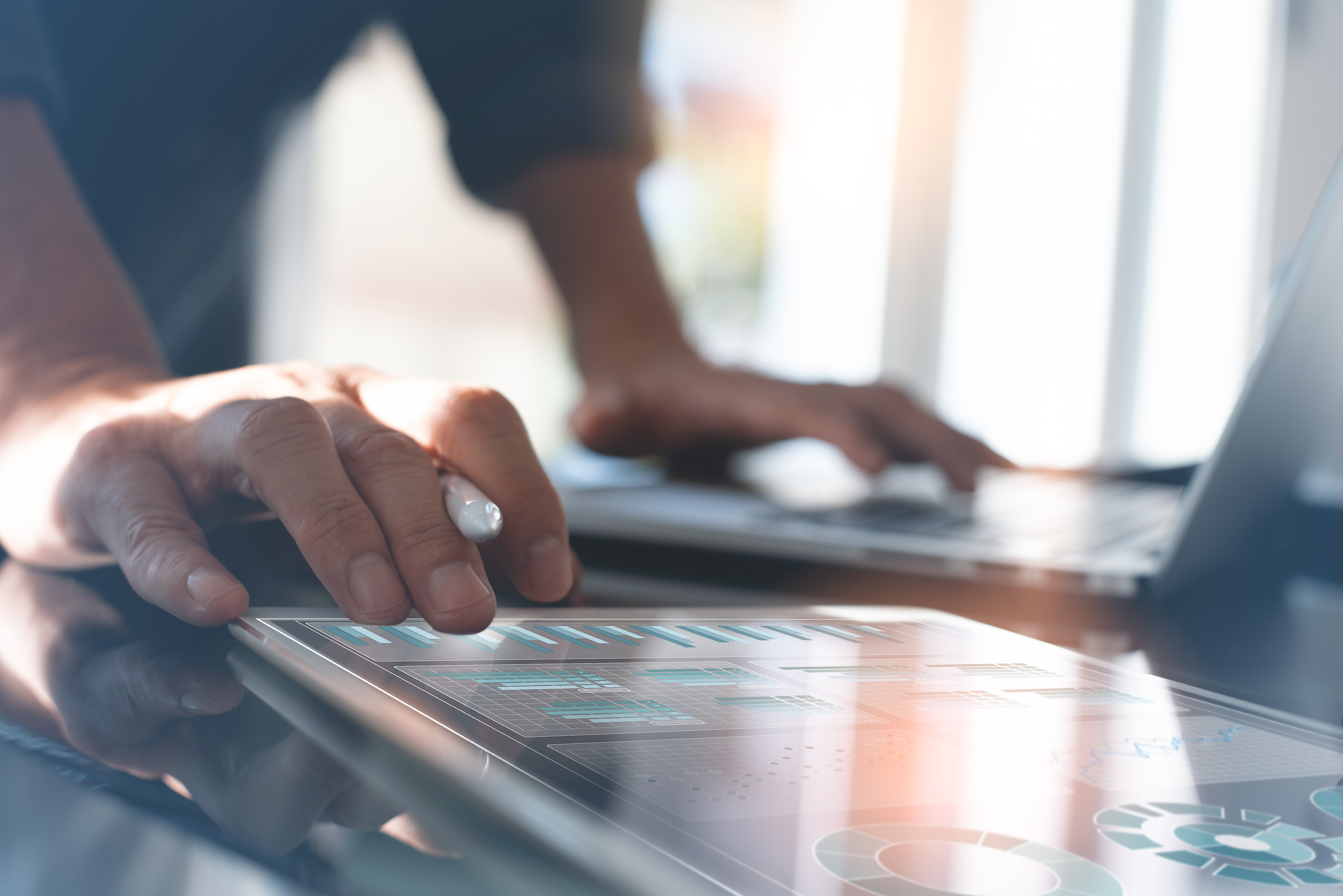 Close-up of a person using a stylus on a tablet displaying data analytics dashboards, with the other hand on a laptop in a modern office environment.