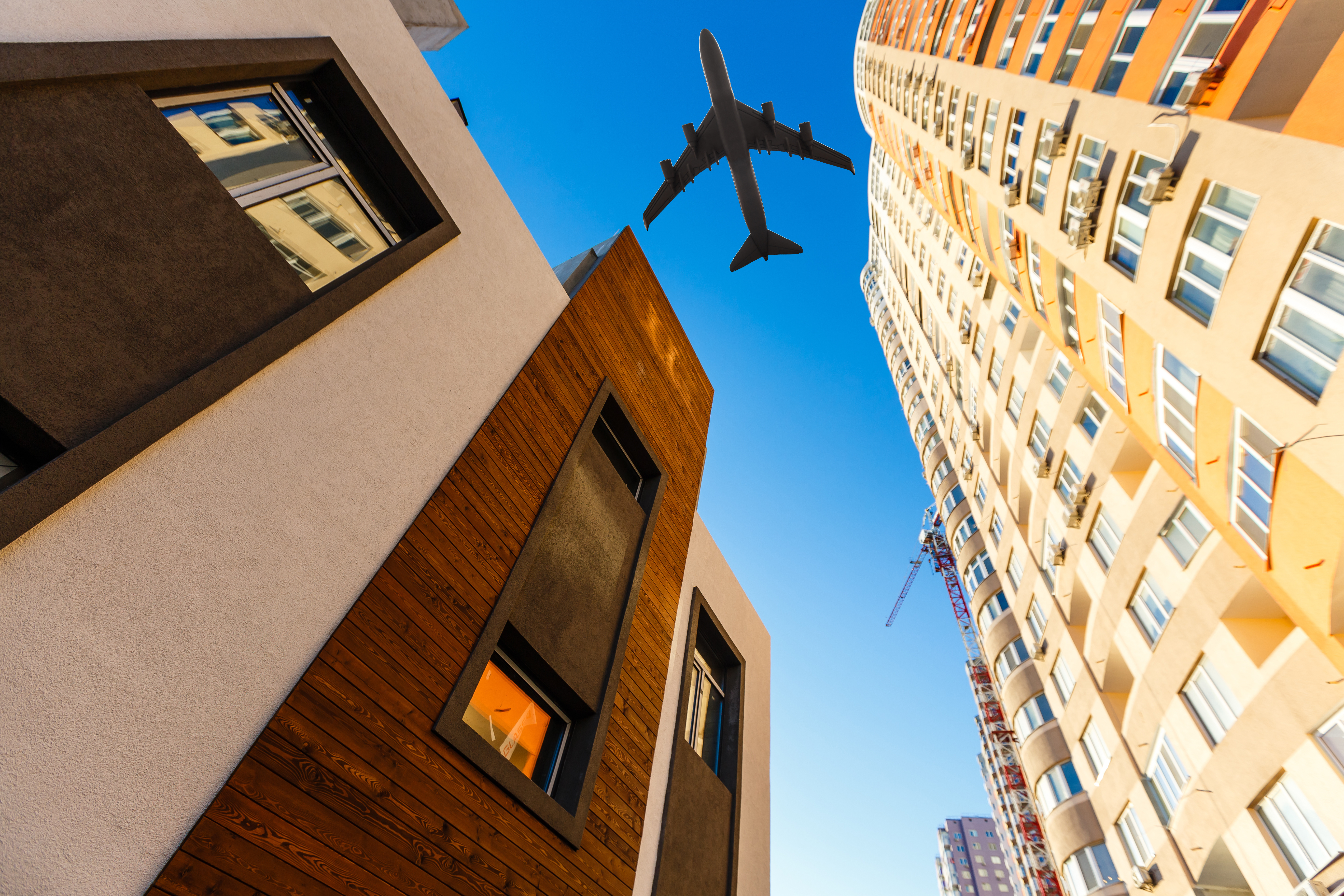 A low-angle view of an airplane flying above modern skyscrapers with a clear blue sky and sunlight illuminating the buildings.