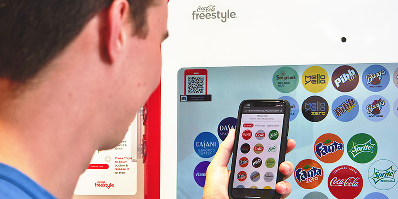 A person selects a beverage on a Coca-Cola Freestyle vending machine using a mobile app, with various drink options displayed on the machine's screen.
