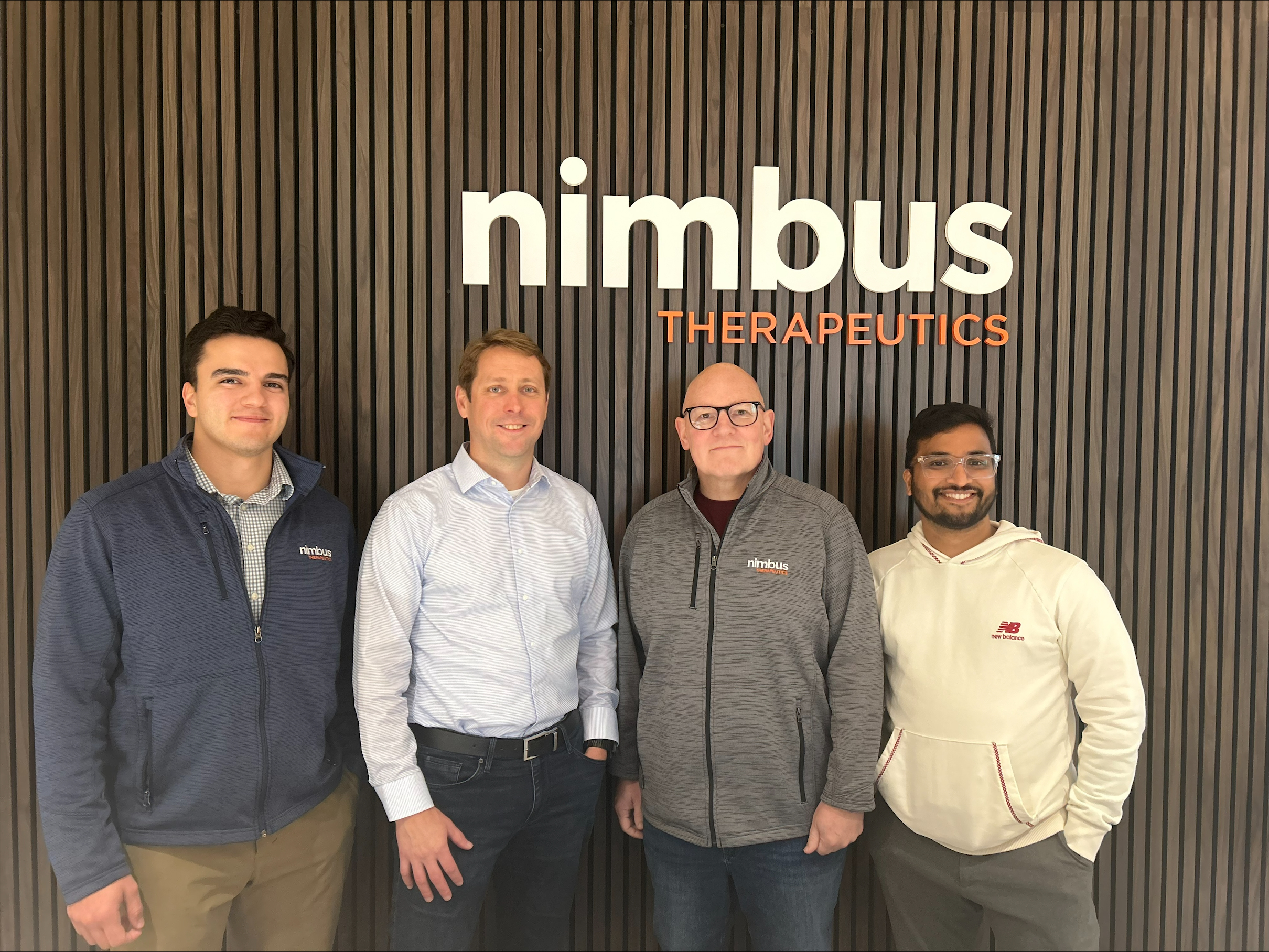 Four members of the Nimbus Therapeutics team standing in front of the Nimbus Therapeutics logo on a wood-paneled wall.
