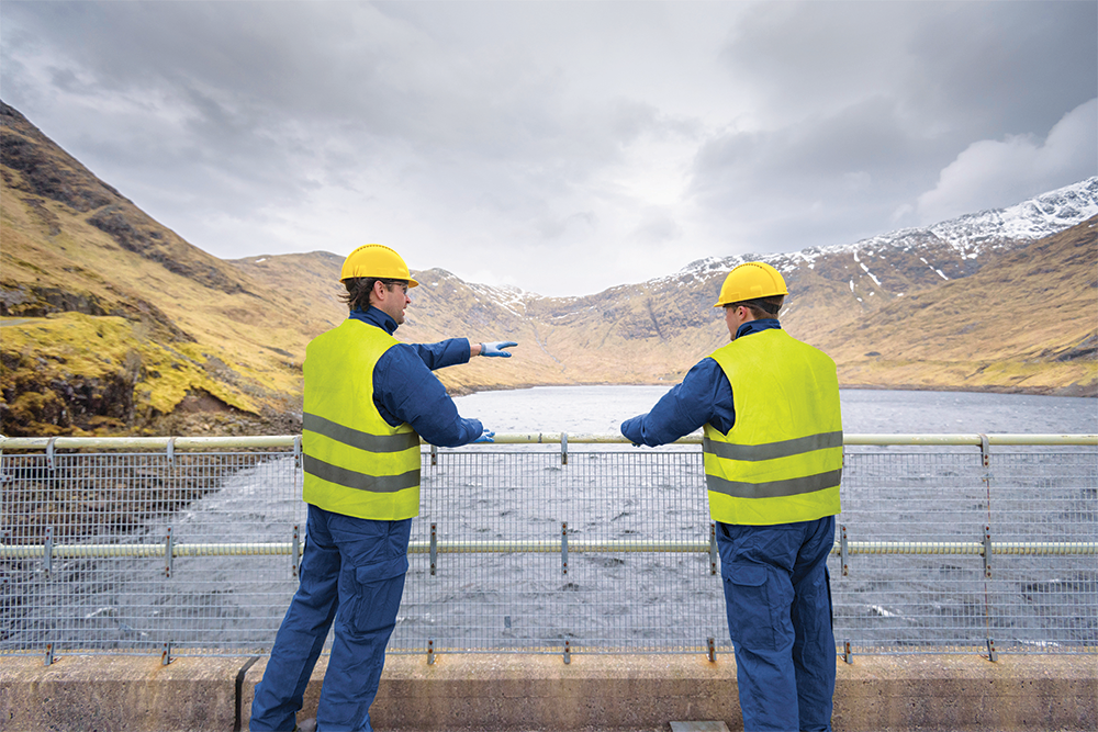 Two engineers wearing yellow safety vests and helmets stand on a dam, overlooking a reservoir surrounded by mountains. One engineer gestures toward the water as they discuss the site, under a cloudy sky.