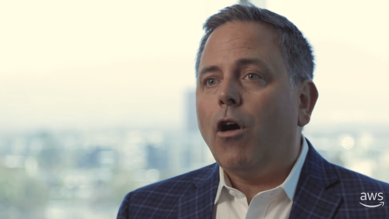 A business professional in a checkered suit speaks during an interview in front of a blurred cityscape background. The AWS logo is visible in the lower right corner.