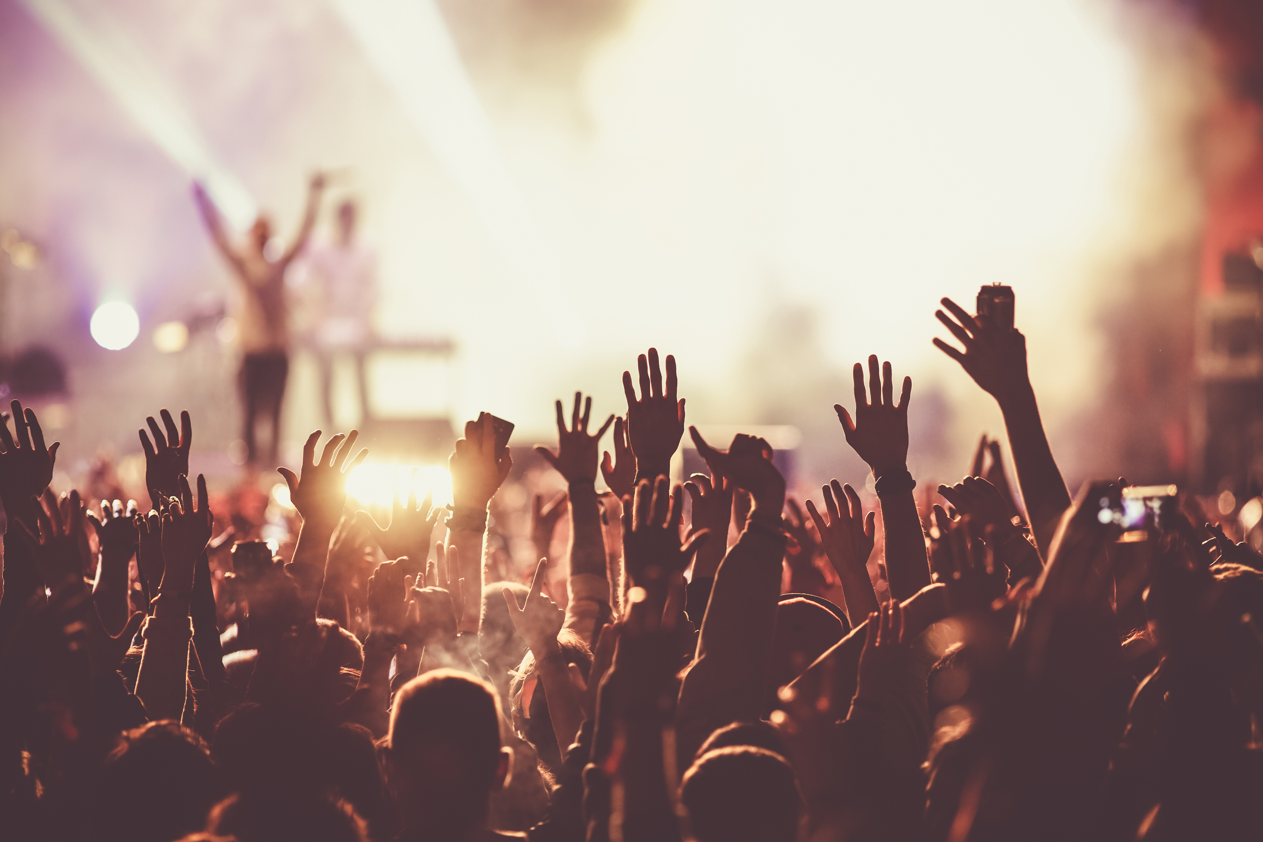 A large crowd of people raising their hands at a live concert or music event, with a brightly lit stage and performers in the background.