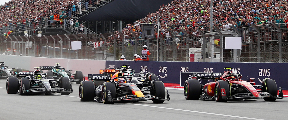 Formula 1 cars racing side by side at the start of a Grand Prix, with a large crowd in the stands and AWS branding visible along the track barriers.