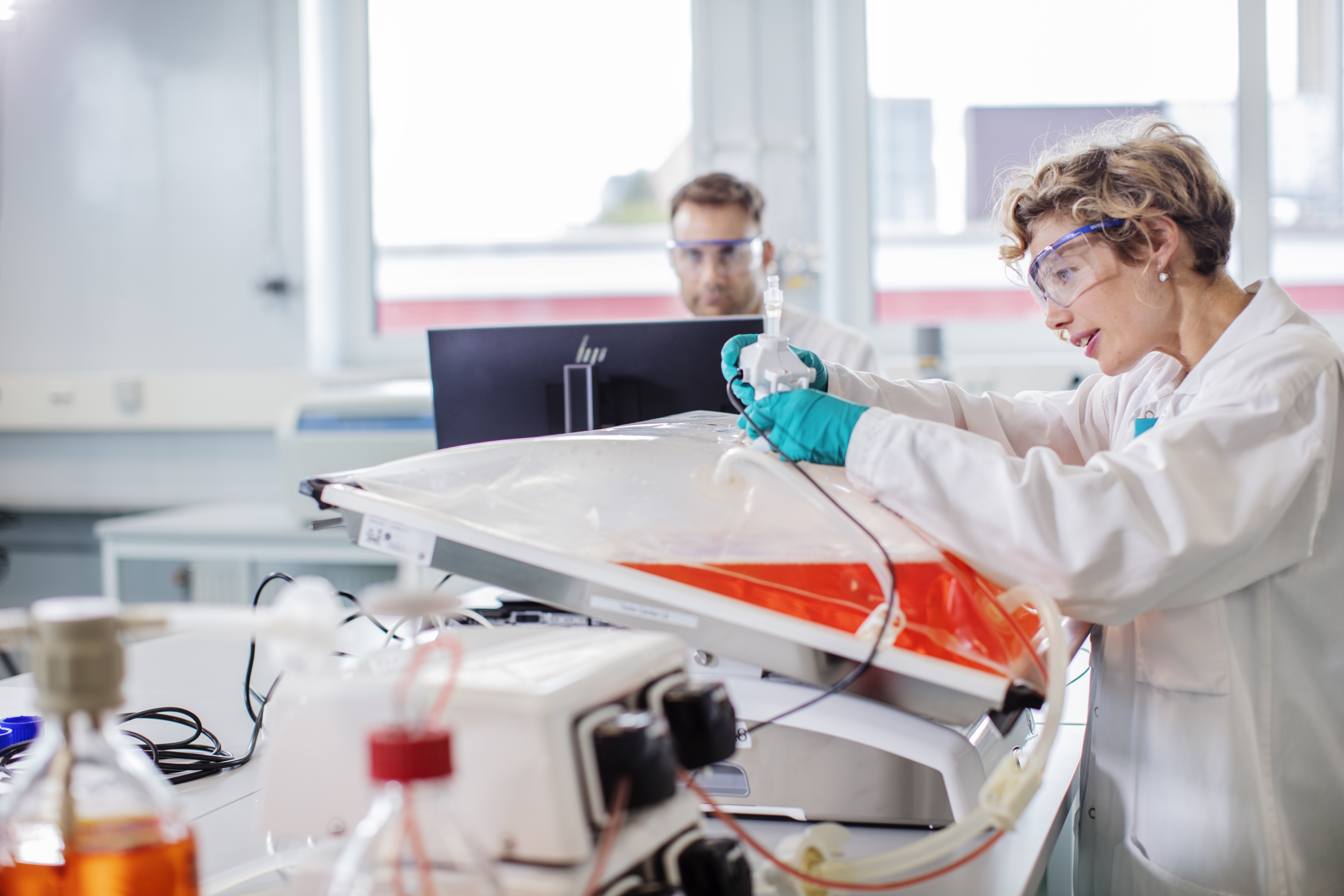 A scientist in a lab coat and safety glasses works with laboratory equipment in a healthcare lab setting, while another person works at a computer in the background.