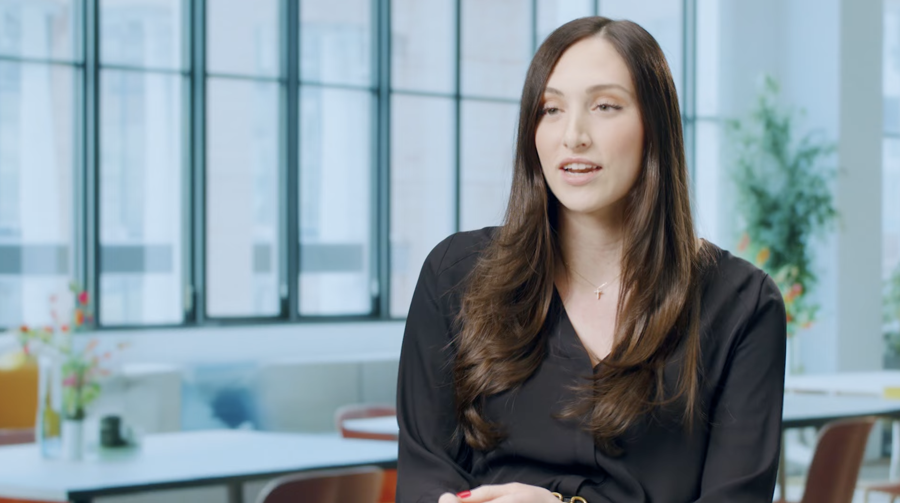 A person with long brown hair wearing a black blouse and a cross necklace, seated in a bright room with large windows and modern furniture.