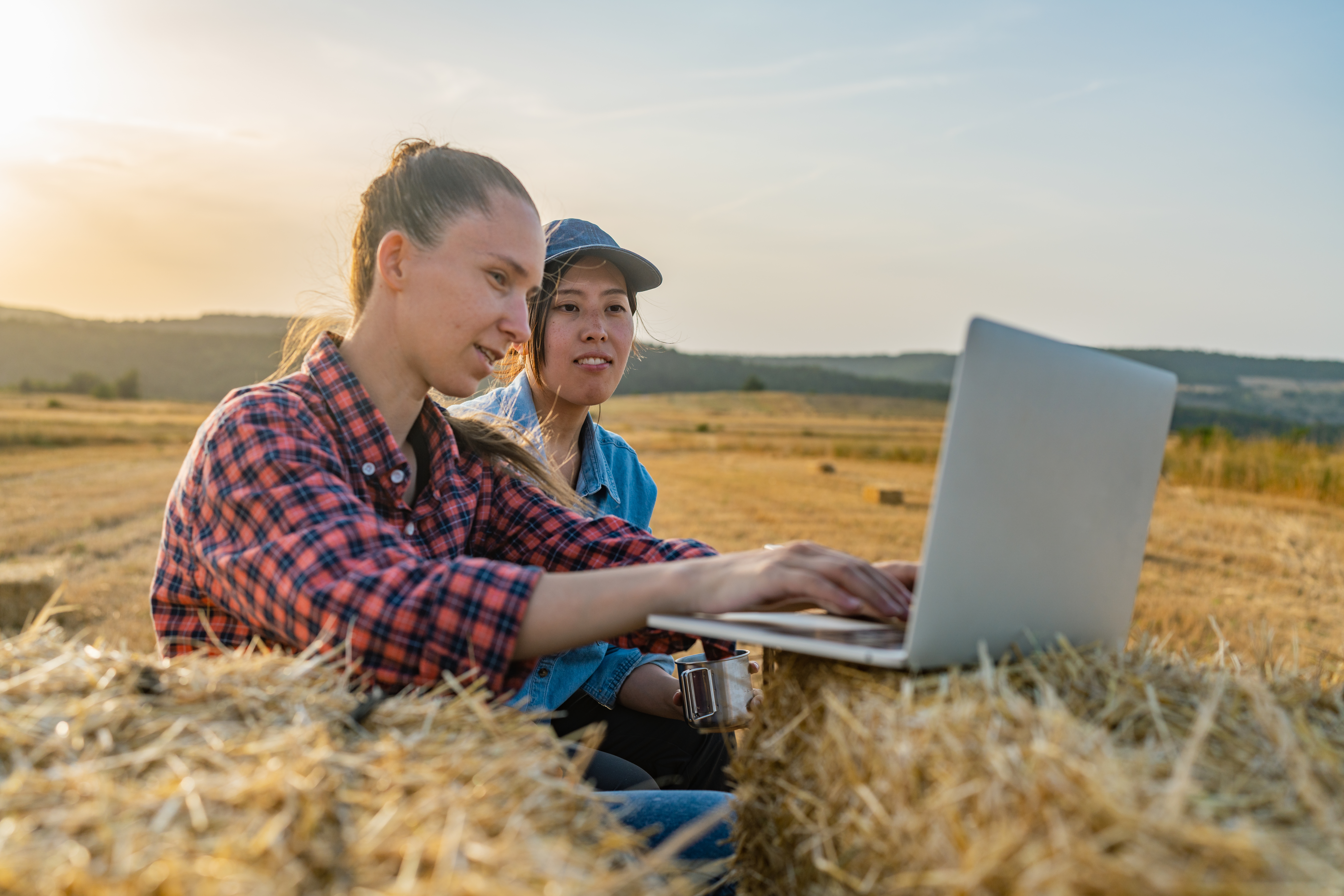 Two female farmers are sitting on a tractor trailer on haystacks and working on a laptop together. Smart farming.