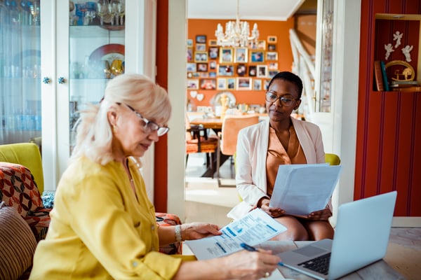 Two women sitting at a table having a financial consultation at home, one holding documents and the other using a laptop, with paperwork and household decor visible in the background.