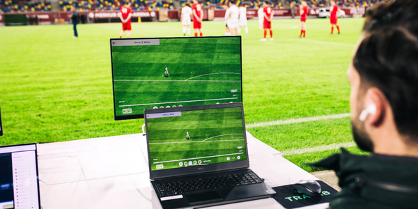 A man operates a TRACAB sports analytics system on a laptop and monitor beside a soccer field during a live match, with players visible in the background.