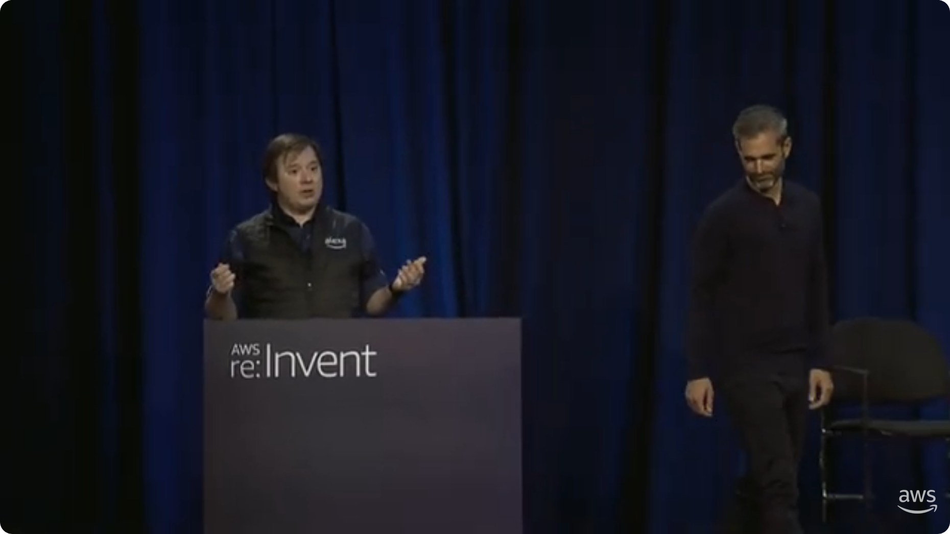 Screenshot of a presentation at AWS re:Invent 2023 featuring two speakers on stage discussing the BMW Group's in-vehicle voice experience project. The speakers are standing on a stage with a podium labeled 'AWS re:Invent' and a dark blue curtain background.