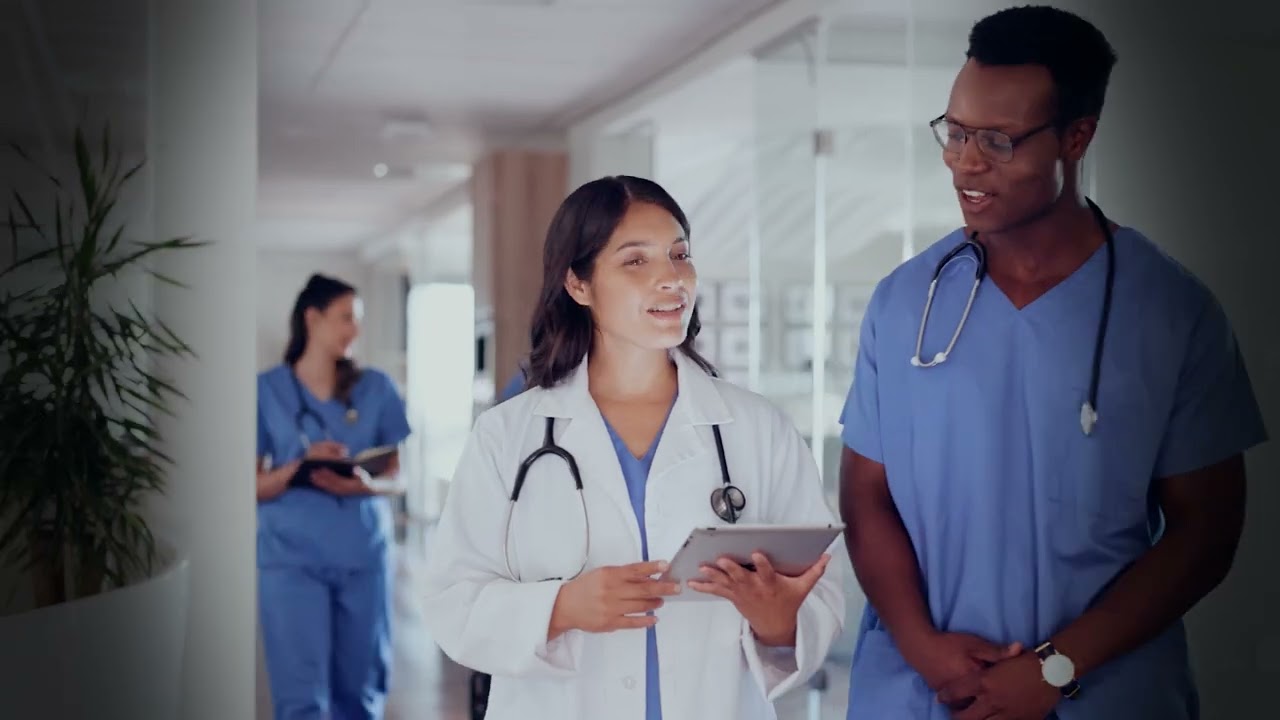 A group of healthcare professionals, including doctors and nurses, discussing and reviewing information on a digital tablet in a hospital corridor. The image conveys teamwork and modern medical practice.