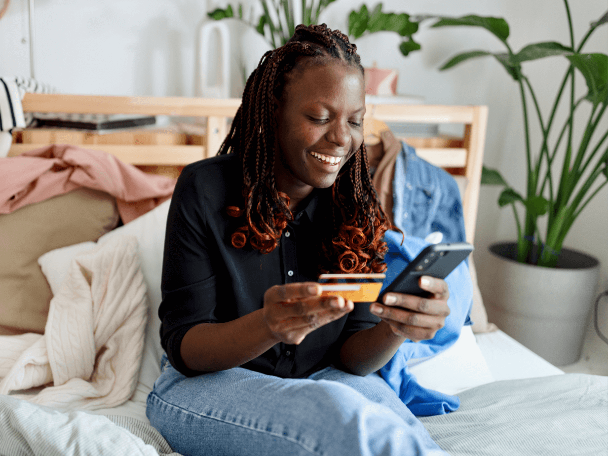 A woman sits on a bed smiling while holding a credit card and a smartphone, making an online purchase in a comfortable home setting.