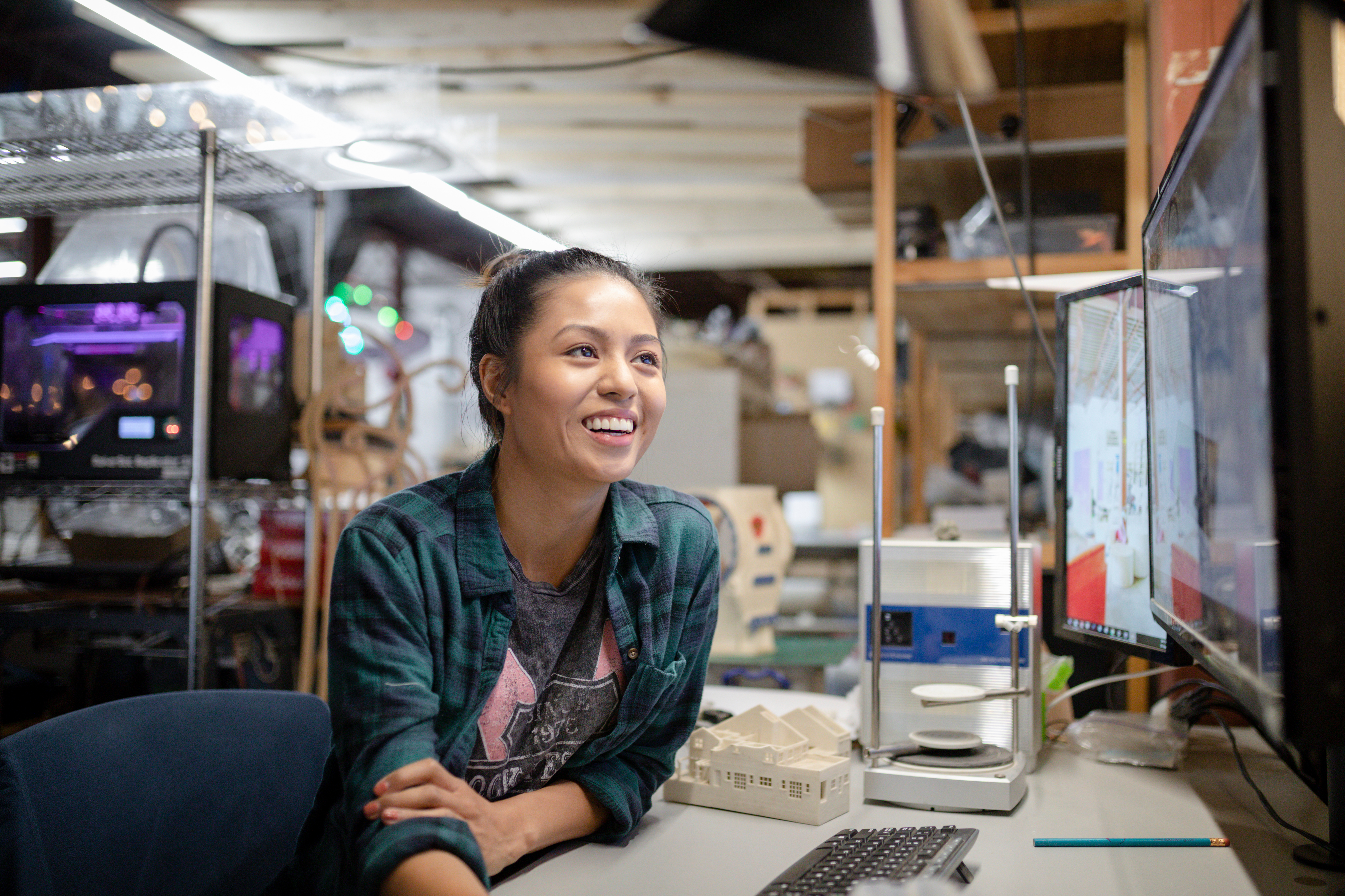 A female engineer smiles while examining a prototype model at her workstation in a workshop, with a computer monitor and 3D printer in the background.