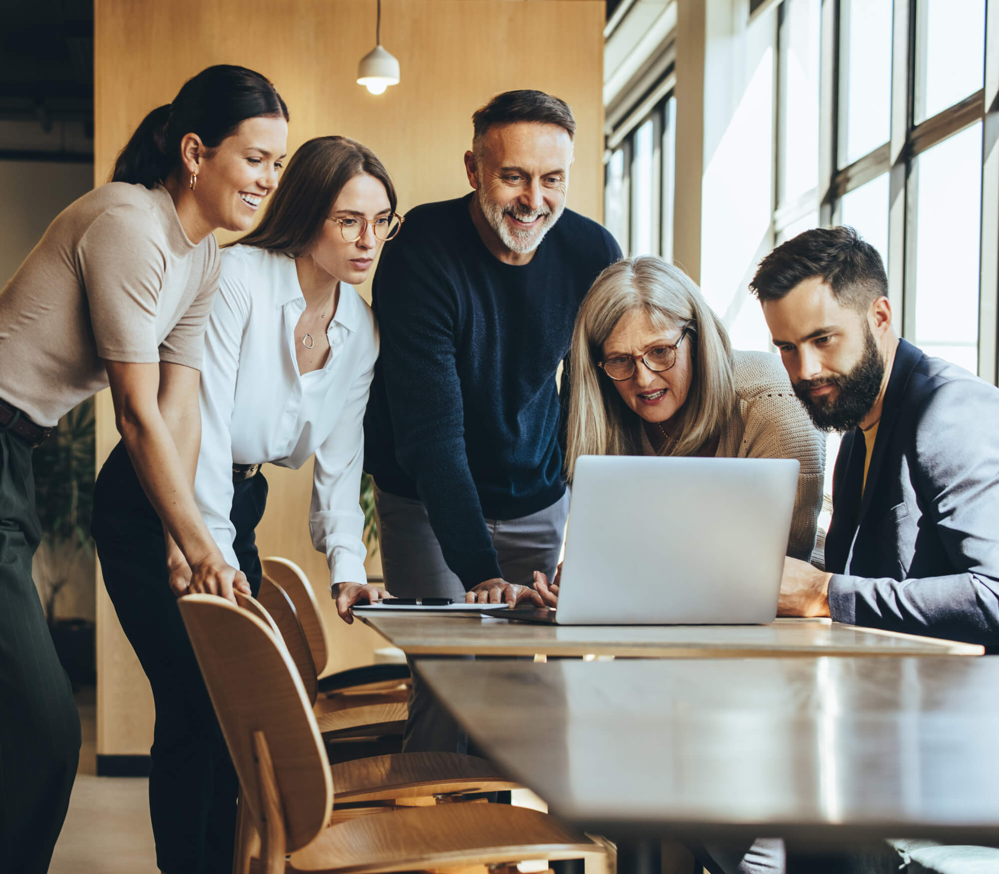 A diverse group of professionals collaborates around a laptop in a modern office space, engaged in teamwork and discussion.