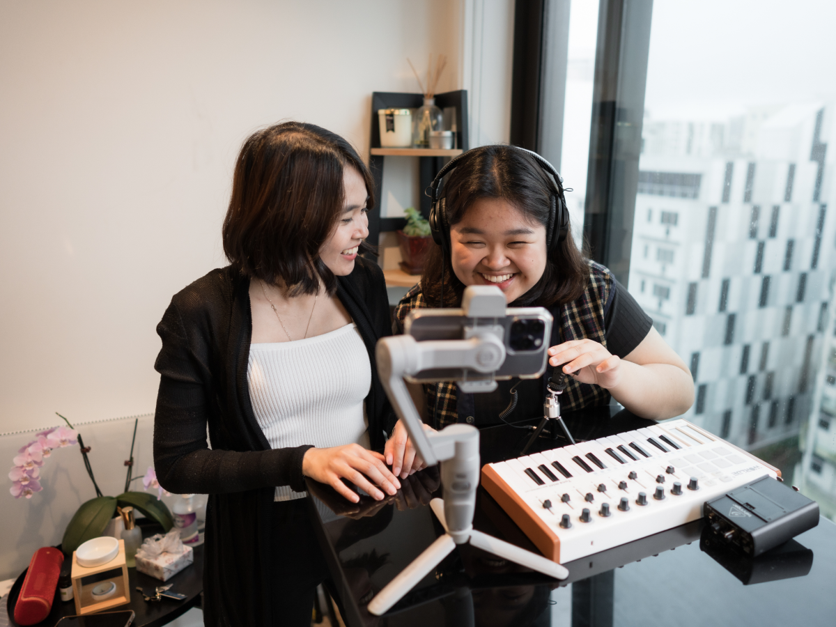 Two women work together to record music using a MIDI keyboard and a smartphone, with one woman wearing headphones and the other assisting, in a modern indoor setting.