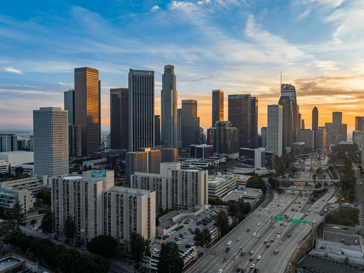 A view of the downtown Los Angeles skyline at sunset, with tall skyscrapers, city buildings, and a busy freeway in the foreground under a partly cloudy sky.
