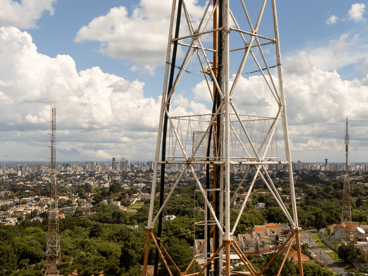Cell phone towers rise above an urban landscape, with a dense city skyline and scattered houses and greenery under a partly cloudy sky.