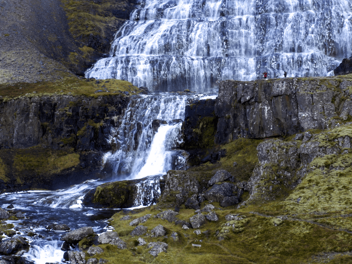 A scenic view of a large waterfall cascading over multiple tiers of rocky cliffs with lush green moss, surrounded by rugged landscape. Two small figures stand on a plateau near the waterfall, highlighting the scale of the natural feature.