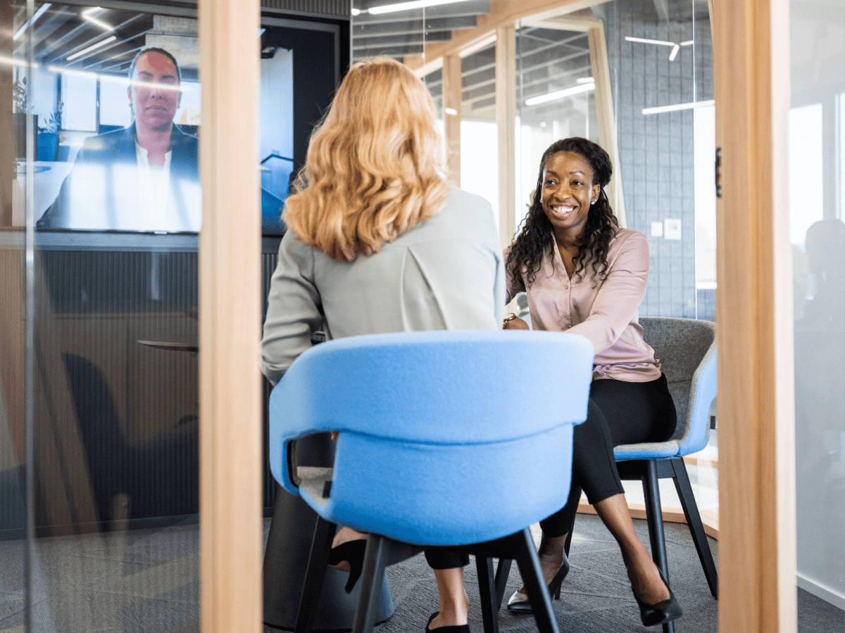 Two women have a business meeting in a modern office conference room, while a third participant joins remotely via video conferencing on a wall screen.