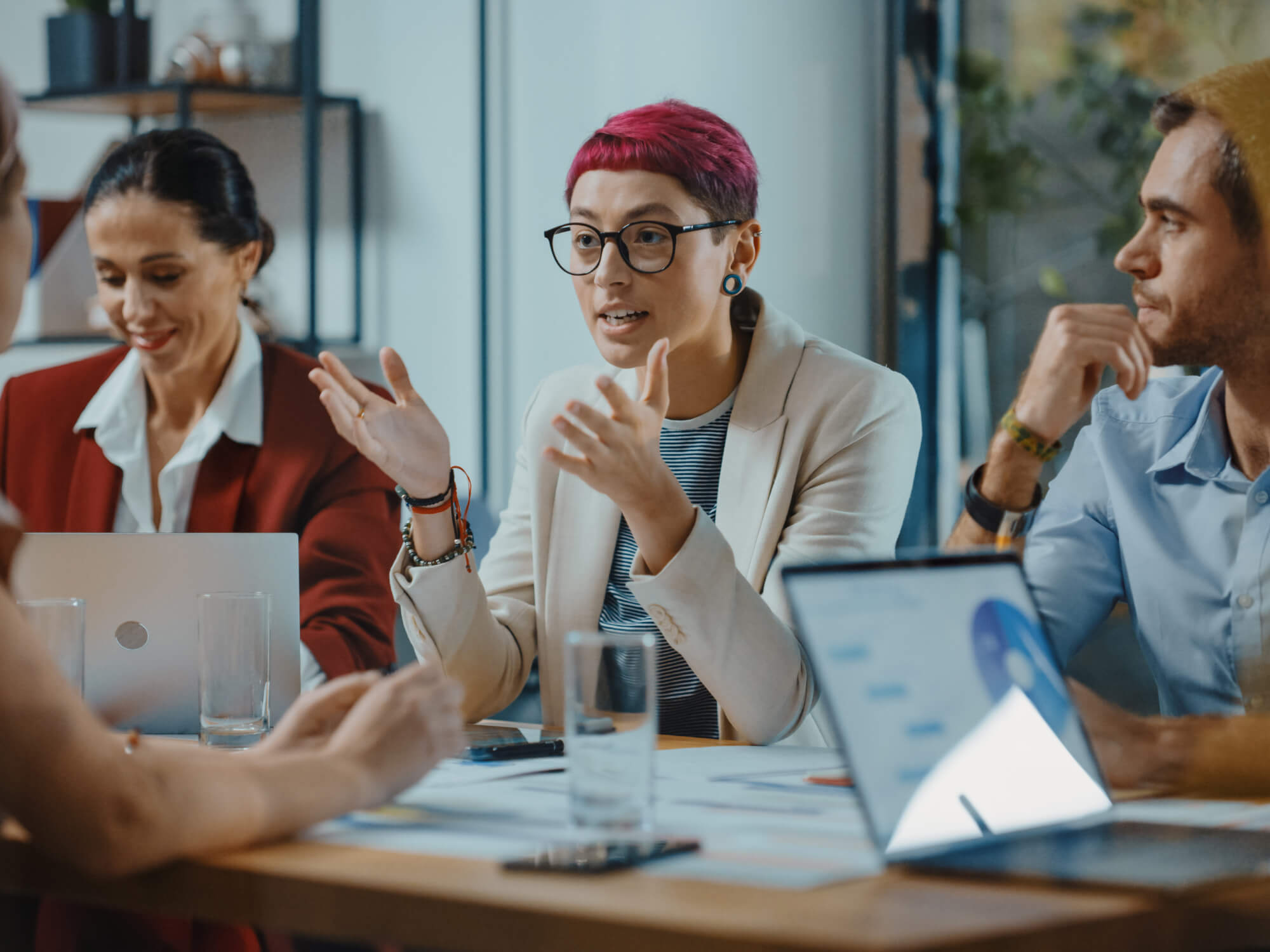 A group of young professionals having a discussion about the benefits of cloud computing in a modern office setting, with laptops and documents on the table.