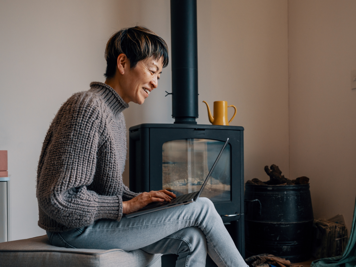 Woman smiling and using a laptop in a cozy room with a wood stove and a yellow teapot in the background.