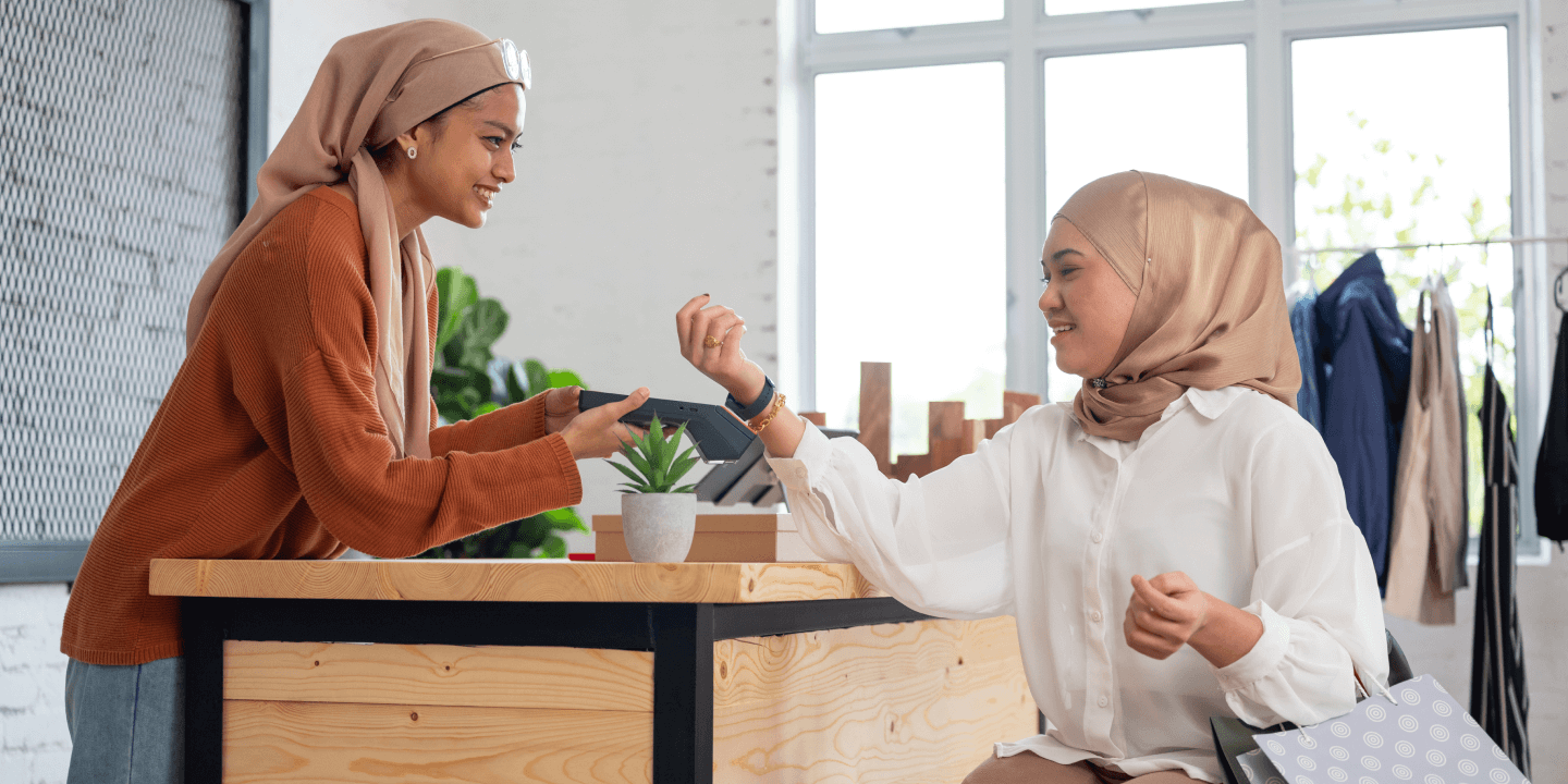 Two women at a retail counter; one is making a contactless payment using a card terminal while the other processes the transaction. Both are smiling and appear to be enjoying the shopping experience.