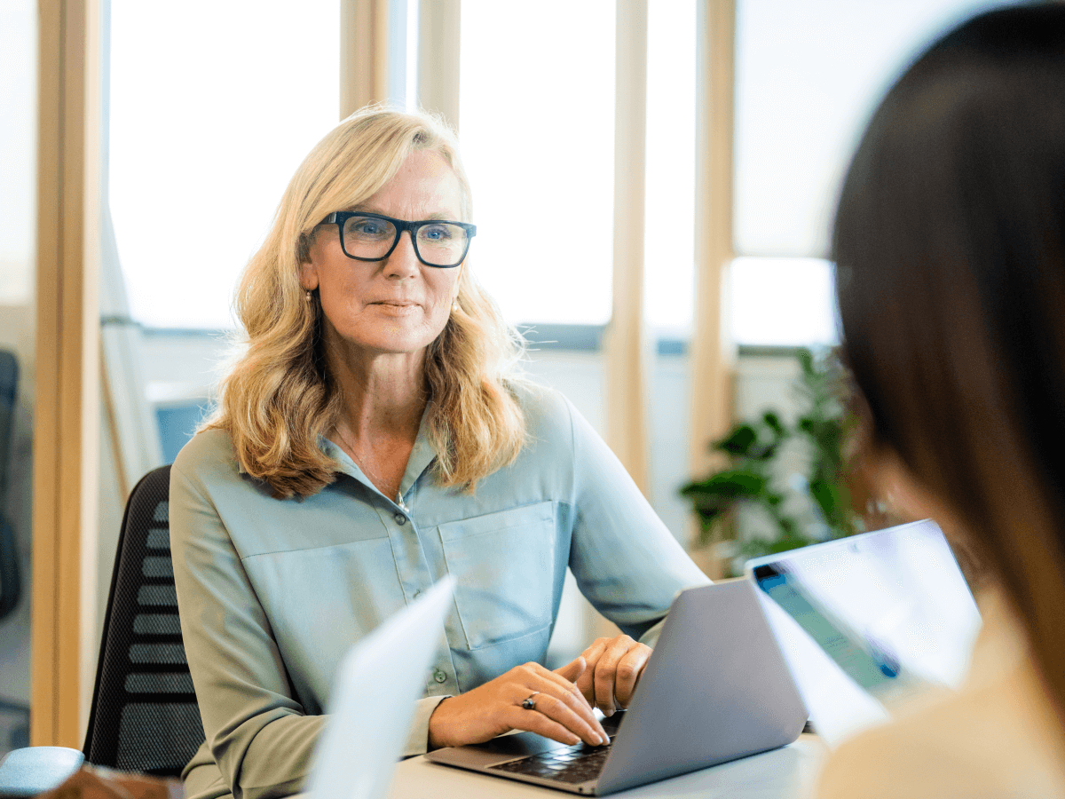 A professional woman with long blonde hair and glasses working on a laptop during an office meeting, engaging in conversation with a colleague.