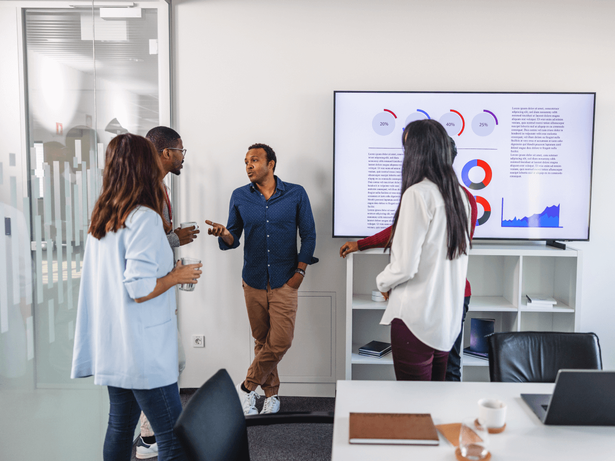 A group of four people engaged in a discussion in an office setting, with a large display screen showing charts and data visualizations in the background.